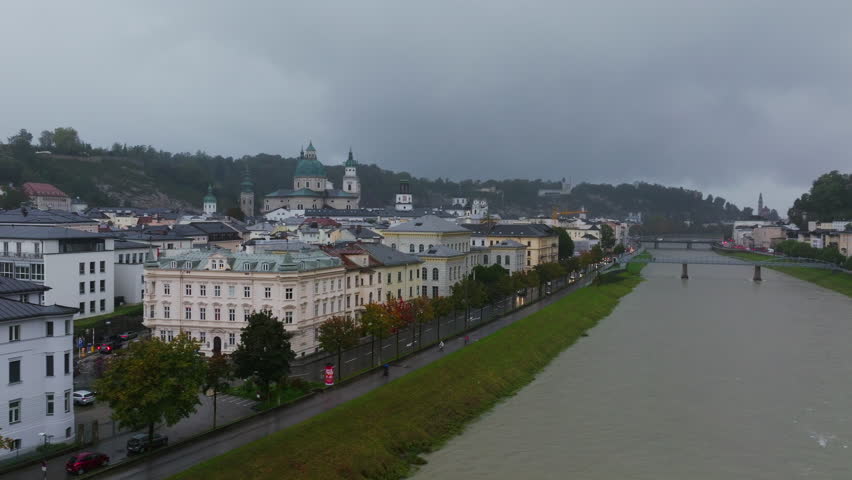 Aerial views showcase Salzburg, Austria, with buildings and houses lining the Salzach River. Cars navigate the roads under a gray, rainy sky, creating a dynamic urban scene