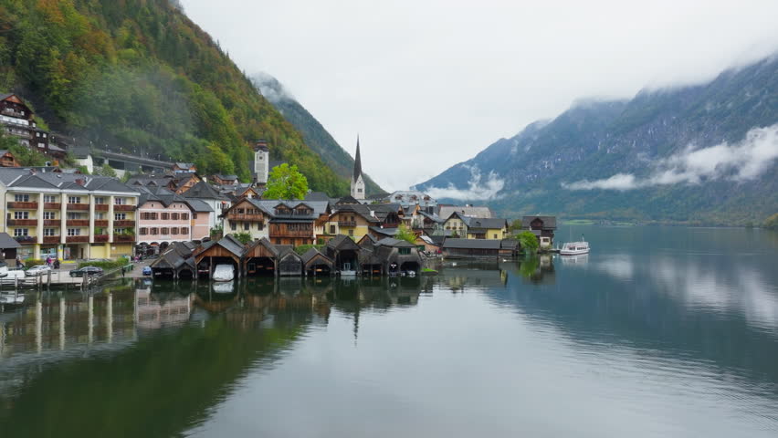 Tranquil Hallstatt, Austria showcases vibrant houses, a church steeple, and majestic Alps in the background. Boats ferry tourists across the serene lake
