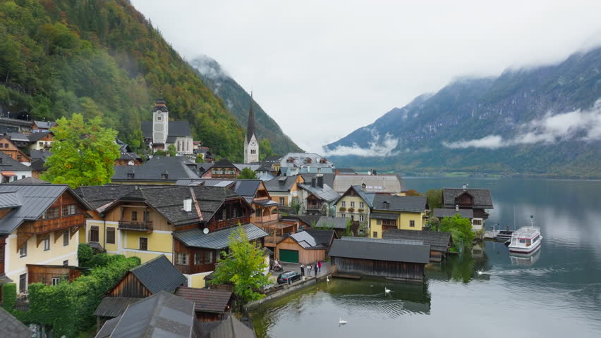 Tranquil Hallstatt, Austria showcases vibrant houses, a church steeple, and majestic Alps in the background. Boats ferry tourists across the serene lake