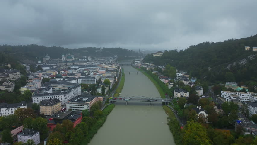 Aerial perspective showcasing the Salzach River winding through Salzburg, Austria. Historic buildings and bridges fill the cityscape beneath a cloudy sky