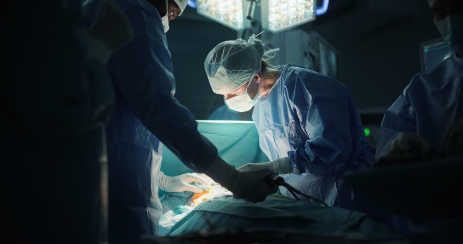 Diverse Surgical Team Working in a Darkened Operating Room Surrounded by Advanced Medical Equipment. Doctors Performing a Complex Surgery in Order to Ensure the Patient’s Safety