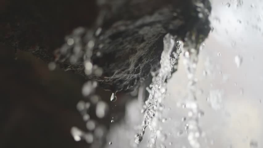Clear droplets drip from rocky ledges in slow motion, each one distinct. Behind, a spring waterfall flows, representing peace and water scarcity issues.