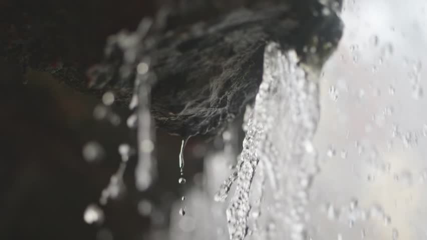 Clear droplets drip from rocky ledges in slow motion, each one distinct. Behind, a spring waterfall flows, representing peace and water scarcity issues.