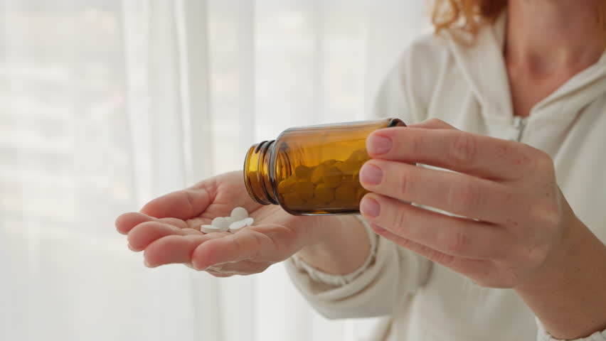 Close-up woman hands pouring white pills from brown bottle. Suggests healthcare, medicine, or supplements, highlighting daily wellness routines and personal health care. Vitamins for women's health