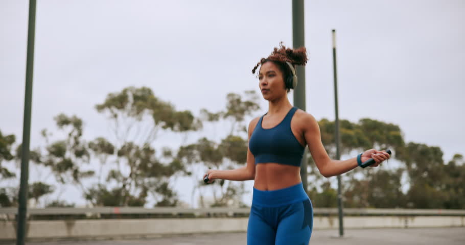 Training, cardio and headphones with woman and skipping rope in city for workout, streaming podcast or challenge. Health, energy and exercise performance with person jumping in town for wellness