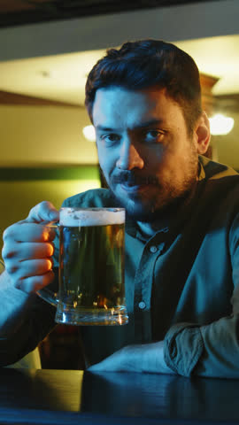 Vertical lowkey portrait of flirty Caucasian man with moustache drinking beer and winking at camera while sitting at bar counter at night