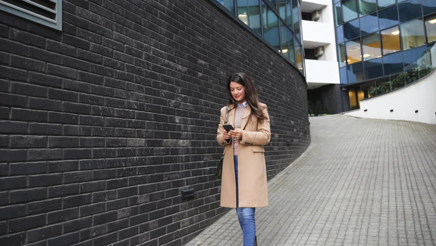 Happy businesswoman texting on smartphone while leaving office building workplace, walking on the city street. Female entrepreneur using mobile phone on the move in the downtown district after work.