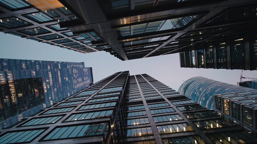 Wide angle gimbal dolly shot downtown high-rise glass skyscrapers in evening, financial district looking up tall modern architecture office buildings city center street, warm lighted windows