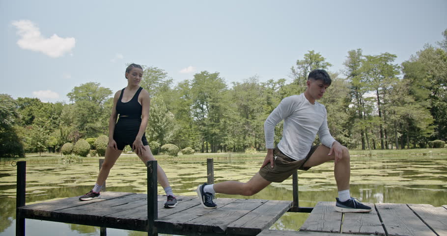 A fit young couple wearing athletic clothing stretches on a wooden dock by a lake on a sunny day.