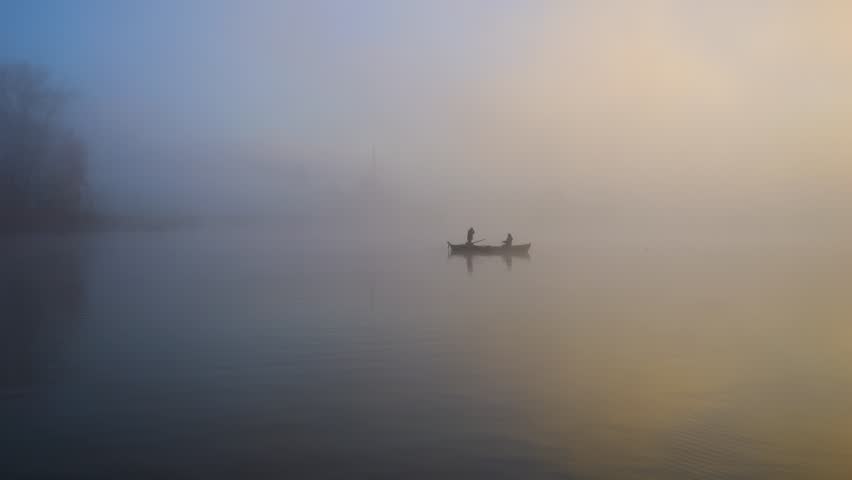Fishermen silhouette are on a fishing boat at sunrise. There is a mystical atmosphere as it is very foggy. The fog covers the landscape and the lake is still. The sun can