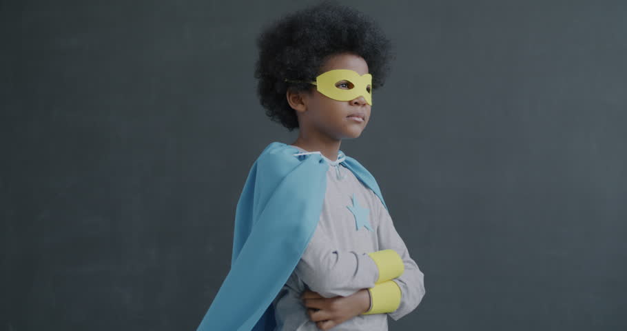 Slow motion portrait of small African American child dressed-up in superhero costume standing on gray background with arms crossed looking at camera with serious face