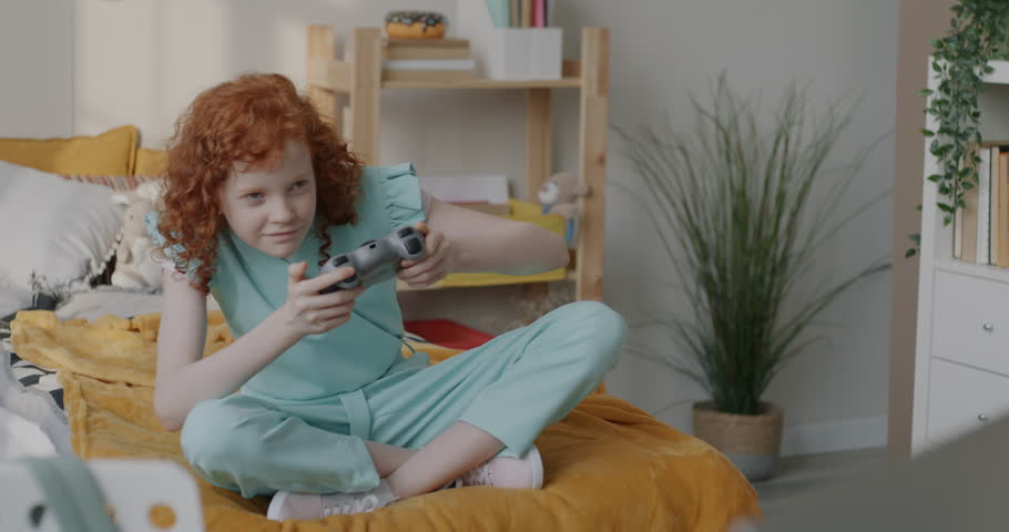 Cheerful girl playing online video game with wireless joystick sitting on bed involved in funny activity at home. Gaming and childhood concept.