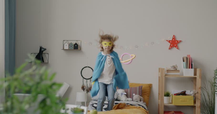 Slow motion portrait of happy kid dressed-up in superhero costume jumping on bed enjoying leisure activity in bedroom. Childhood and fun concept.