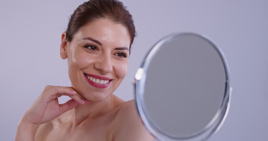 Young brunette cheerful pretty woman with natural make up and beautiful fresh clean skin looking in the mirror and smiling isolated on grey background . Skin care concept. 4k video.