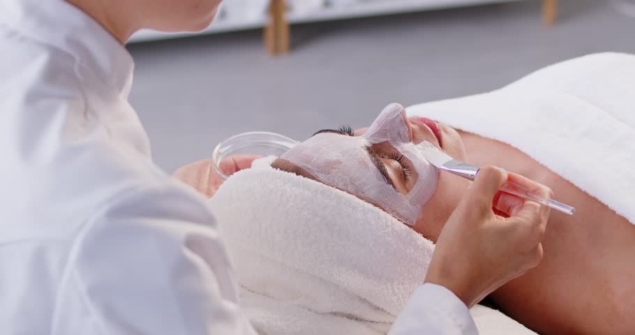 Dermatologist, cosmetologist applying skin care cream with brush on female face, mask for luxe feeling, moisturizer, beauty professional hygienic relaxing session in spa salon or beauty clinic