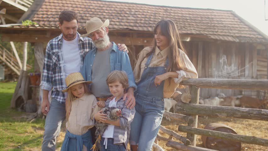 Positive Caucasian family gathering together. Standing in front of barn. Looking at camera while smiling. Little boy holding chicken with both hands. Girl trying petting animal. Gathering.