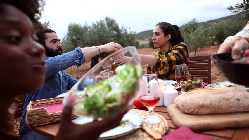 Multi ethnic group of farmers are having dinner together at a wooden table in an olive grove, sharing food and drinks as the sun sets, creating a warm and convivial atmosphere