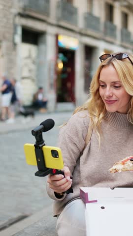 Vertical video of a young woman savoring a slice of pizza while vlogging on a bustling city street, capturing moments of joy and urban life. Influencer enjoying pizza while vlogging