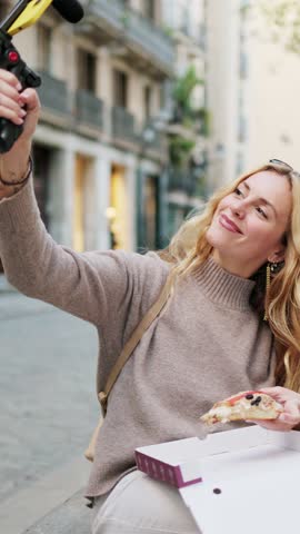 Vertical video of a travel influencer savors a slice of pizza while creating content for her vlog on a bustling city street. Influencer enjoying pizza while vlogging on city street