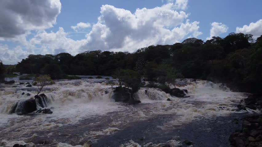 The aerial frame captures fragments of trees and rocks embedded within the river's rapids while the camera ascends, revealing a broader perspective.