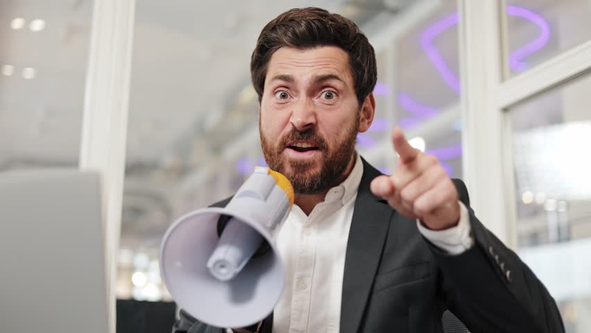 Angry businessman yells into a megaphone and points his finger at camera. Man is sitting at desk in office environment. Annoyed male in formal clothes speaking in megaphone and making hand gestures. .