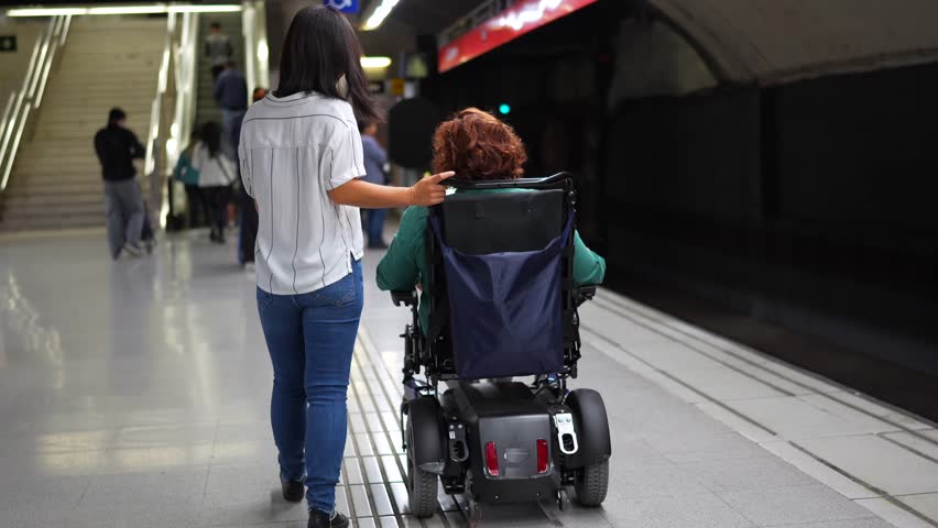 Rear view of a disabled caucasian adult woman in wheelchair and asian friend walking along metro station
