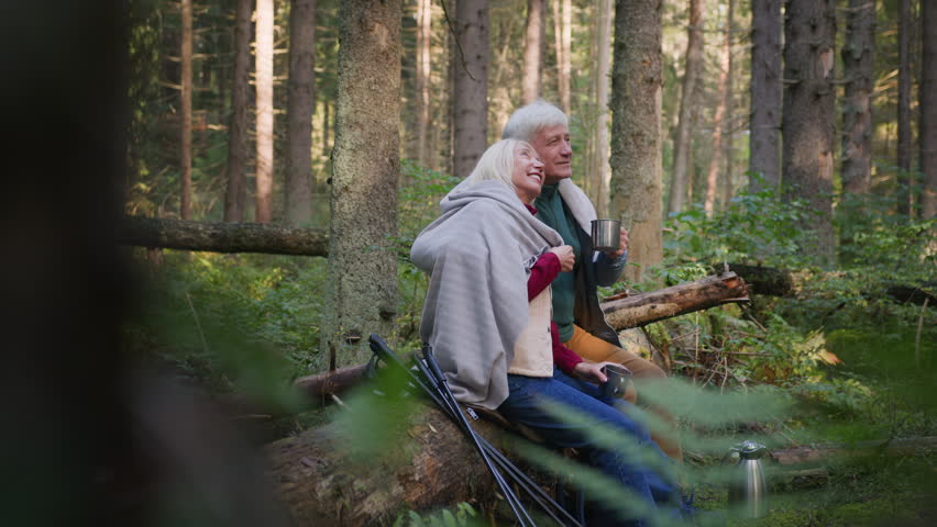 Senior couple enjoy walking in forest on sunny day. Elderly positive man and woman sit together for hot drink and rest, admiring nature. Old husband cover wife with plaid and hug close to warm up