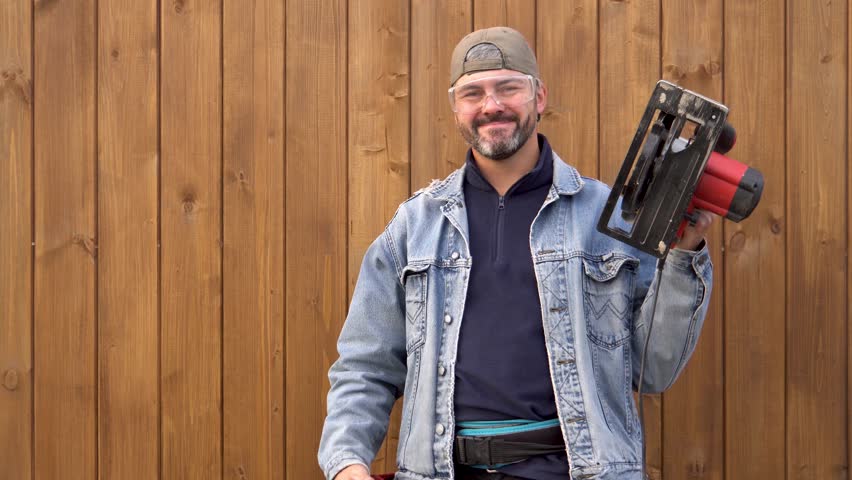 Adult man holding electric saw. Circular saw. Construction tool. Builder. Vertical boards on background. Village sil. Farmer. Worker. Countryside. Outdoors. Close-up.