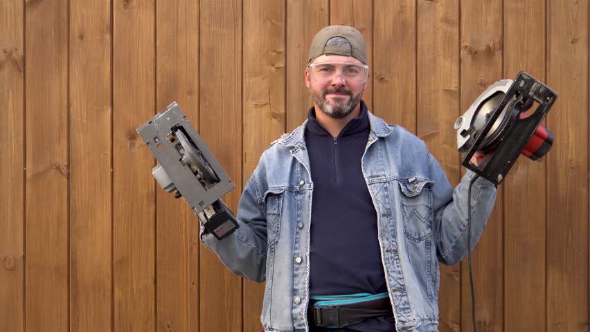 A man poses with electric saws in his hands. Stands against a wooden wall. Bearded. Dressed in a denim jacket. Daylight. Builder. Worker. Master. Close-up. Portrait.