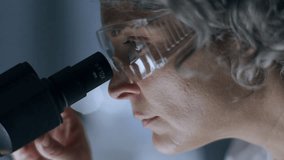 Close-up view of grey-haired female scientist wearing protective goggles, looking through microscope binoculars, performing research in laboratory. Close-up view - Powered by Shutterstock - Get 15% off with code: PIKWIZARD15