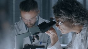 Middle-aged female scientist with grey hair, wearing protective glasses, examining sample through microscope and taking notes, as her male colleague working in background in laboratory - Powered by Shutterstock - Get 15% off with code: PIKWIZARD15