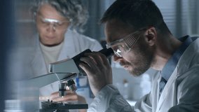 Male scientist in safety eyewear observing sample under microscope and writing down notes, as his female colleague working beside at desk in laboratory - Powered by Shutterstock - Get 15% off with code: PIKWIZARD15