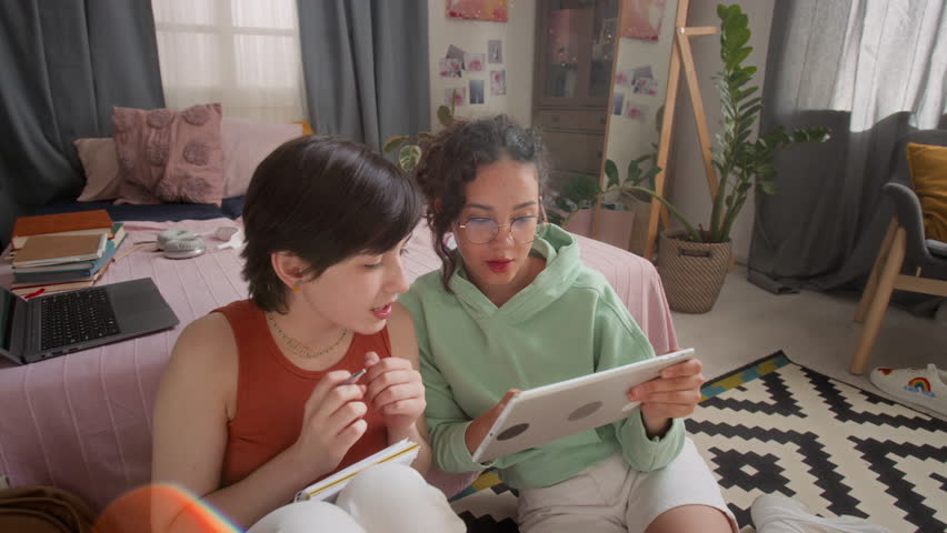 Two teenage girls sitting closely together on floor in cozy, sunlit room, looking at digital tablet screen, talking and clapping hands
