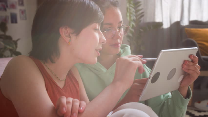 Two girls, sitting closely on floor in cozy sunlit bedroom, looking at digital tablet, smiling and chatting while scrolling through social media
