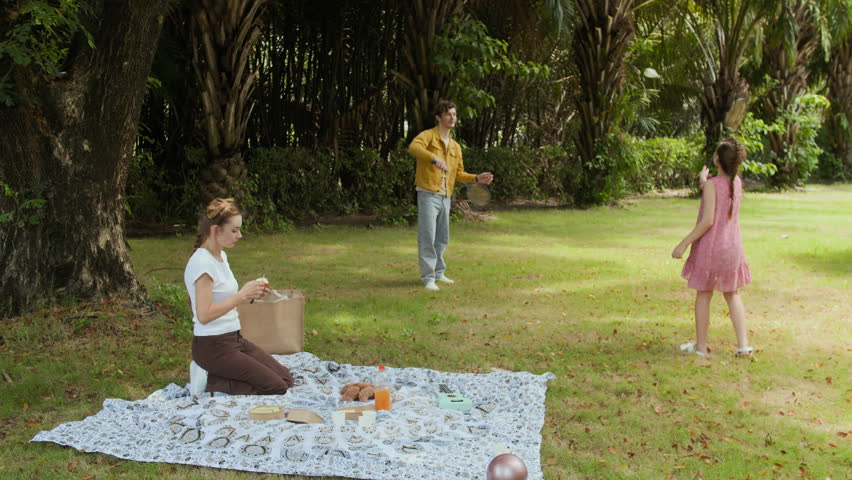 Very long shot of father and daughter playing badminton while mother serving snacks on blanket during family picnic in park