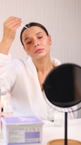 happy woman applying serum or cream on face, during facial ritual at home.smiling girl with natural skin using massage roller.double side mirror and cosmetic jars in front,monstera leaf as decor.