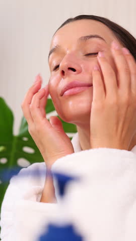 happy woman applying serum or cream on face, during facial ritual at home.smiling girl with natural skin using massage roller.double side mirror and cosmetic jars in front,monstera leaf as decor.