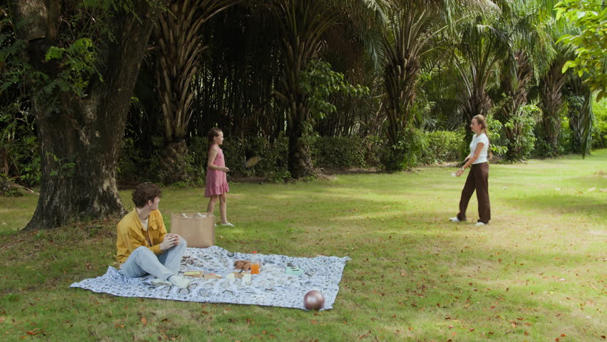 Very long static shot of young mom and daughter playing badminton while dad sitting on blanket during picnic in summer park