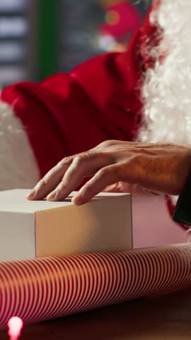 Vertical Video Santa Claus decorates a gift box in his festive workshop preparing wrapped presents and ornaments to fill the holiday season with magic. Christmas delivery from the North Pole. Camera A
