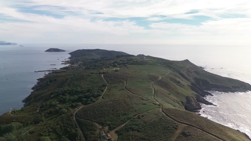 Aerial view over Ons Island, capturing its winding paths, rocky cliffs, trees, lighthouse and Barrio do Curro with harbor pier. Tranquil Atlantic Ocean and cloudy skies. Onza island in background.