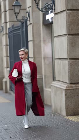 Stylish Woman in Trendy Burgundy Coat Walks City Street, Wearing Fashionable clothing White Turtleneck Sweater, Pearl Necklace and Grey Jeans, White Bag. Confident and Chic Mature Fashion Model