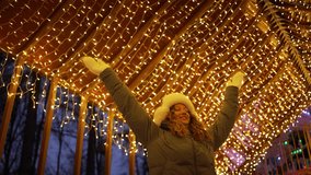 Young woman bundles up for winter, raising her arms in joy while surrounded by the enchanting glow of christmas lights at an amusement park during a festive evening - Powered by Shutterstock - Get 15% off with code: PIKWIZARD15