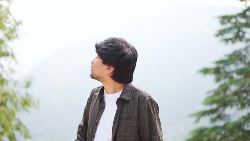 4K Portrait shot of an Indian man wearing casual clothes staring at the sky while being lost in deep thoughts at Manali in Himachal Pradesh, India. Indian man looks at sky searching for hope. 
