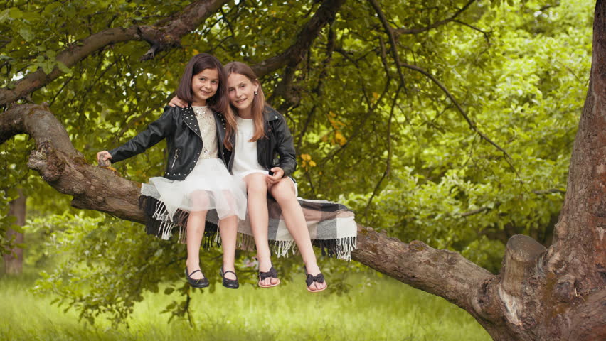 Two little sisters in summer dress and black jacket sitting together on branch of old tree. Cheerful cute girls spending free time at green garden. Summertime and family concept. Little sisters.