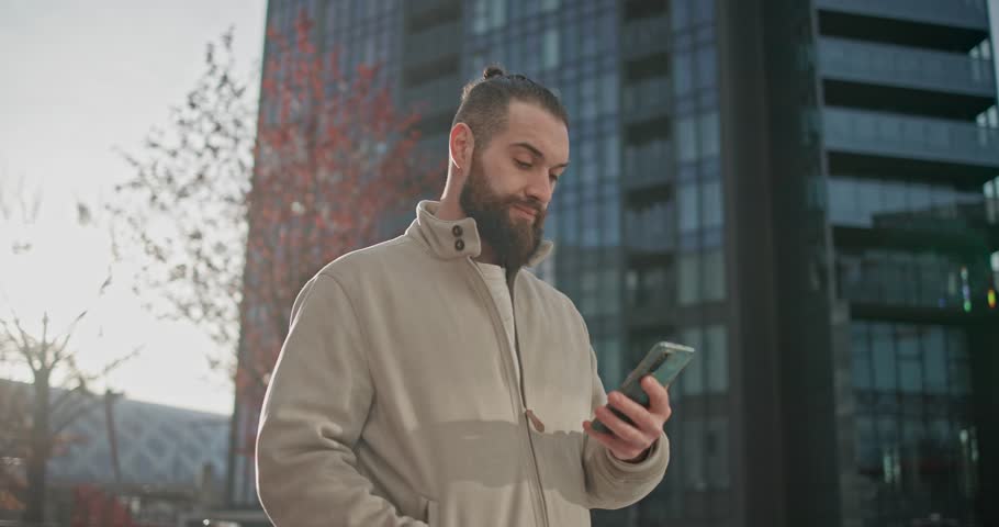 Young man with stylish trendy hairstyle and beard in casual clothes in urban environment standing against modern buildings background and reading messages or news on phone