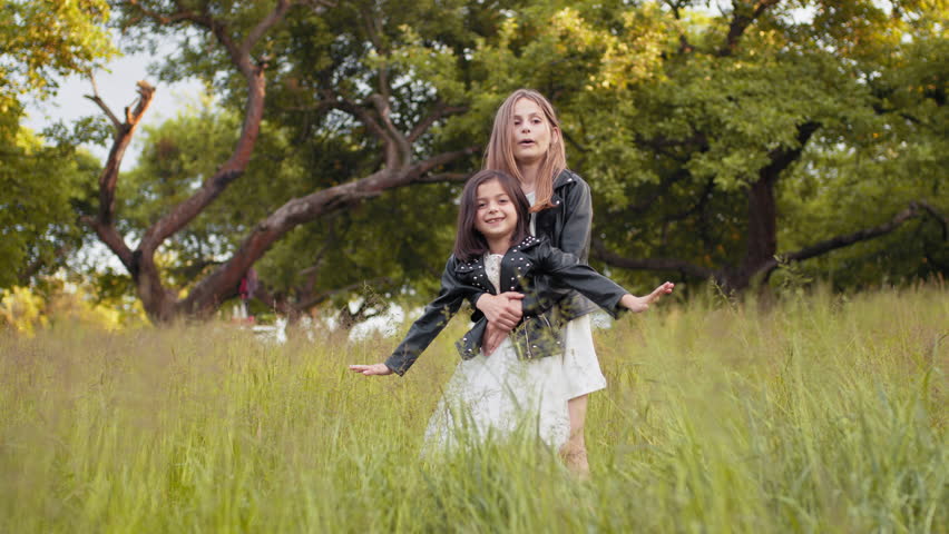 Two caucasian little girls in summer dress and black jacket playing together at summer garden. Pretty sisters in positive mood relaxing on fresh air. Caucasian sisters playing together at green garden