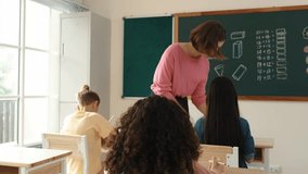 Smart teacher checking cute student homework while explaining idea at class. Attractive instructor walking around classroom while looking at notebook and talking about Math theory to child. Pedagogy. - Powered by Shutterstock - Get 15% off with code: PIKWIZARD15