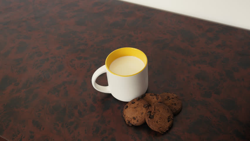 A cup of milk and chocolate chip cookies on the side. A single cookie being dipped into the mug by a female hand. With milk splashing around and a single drop falling of the cookie 