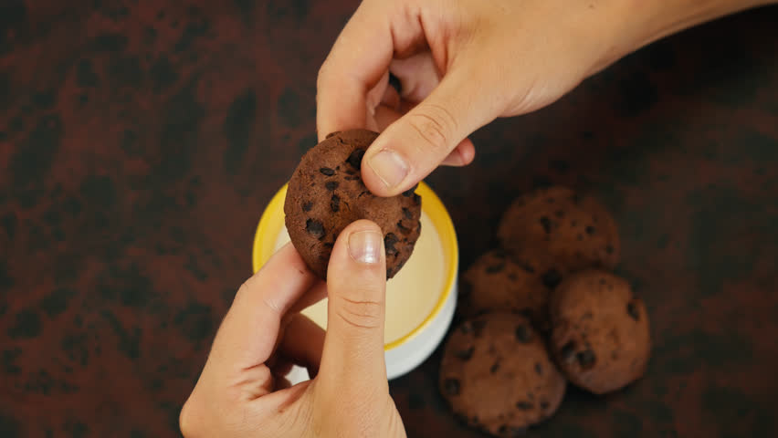 Chocolate chip cookie being cracked into two pieces by female hands over a cup of milk with other biscuits on the side. The crumbs falling into the cup.
