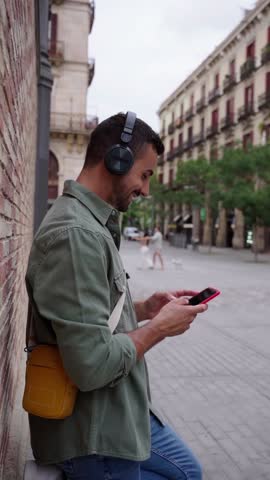 Cheerful caucasian man relaxing outdoors using phone sending text messages and checking the social media and listening music leaning in a brick wall in an urban street. Vertical footage.